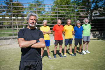 Obraz premium Portrait of serious senior man on football field. Captain with gray hair in sport clothes standing, looking at camera, crossing arms, teammates in background. Football, sport, leisure concept