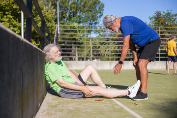 Senior man injured during football training. Caucasian man with grey hair in sport clothes sitting on ground, touching leg, friend helping. Football, sport, leisure concept