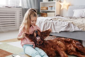 Portrait of cute blonde girl playing with Irish Setter dog on floor in cozy home interior, copy...