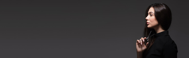 Fototapeta premium side view of woman holding scissors near long shiny hair isolated on dark grey, banner