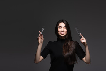 Obraz premium cheerful young woman with long shiny hair holding scissors and comb isolated on dark grey