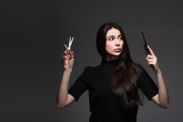 Obraz premium young woman with long shiny hair holding scissors and comb isolated on dark grey