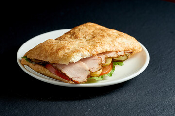 Ciabatta sandwich with tomatoes and boiled pork and feta cheese in a white plate on a black background