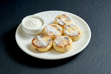 Obraz na płótnie Canvas Cheesecake pancake with sour cream in a white plate on a black background. Syrniki