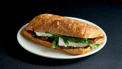 Ciabatta sandwich with baked vegetables and arugula and feta cheese in a white plate on a black background