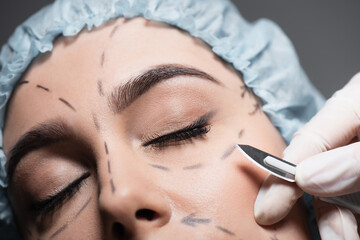 close up of plastic surgeon in latex glove holding scalpel near woman with closed eyes and marked lines on face isolated on grey