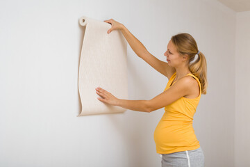 Young adult pregnant woman standing at white wall and looking at new wallpaper for room. Side view....