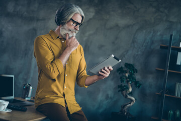 Photo of smart clever mature guy dressed yellow shirt spectacles arm chin reading modern gadget indoors workshop workplace