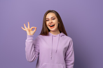 cute smiling brunette girl showing OK sign with happy face over purple background. Looking at camera. Positive emotions concept