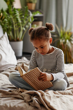 Vertical portrait of cute African-American girl reading book while sitting on bed in cozy interior
