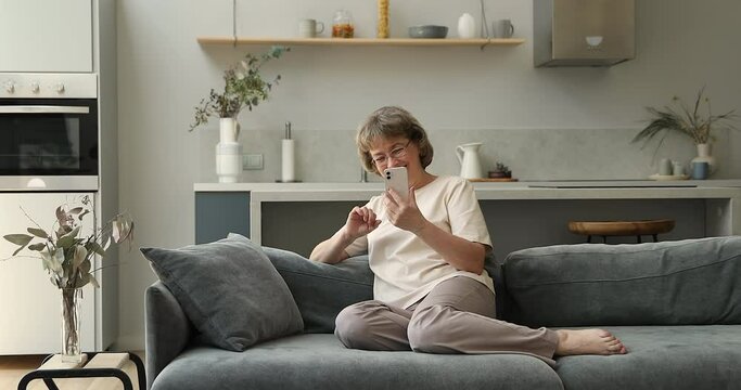 Positive middle aged lady on pension enjoy rest on sofa at home having fun with modern mobile phone test new application scroll social media content. Laughing mature female work online using cellphone