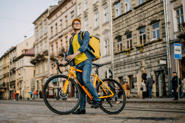 Obraz na płótnie Canvas A happy delivery man who is sitting on a bicycle and looking around. He is carrying a yellow backpack on his shoulders.
