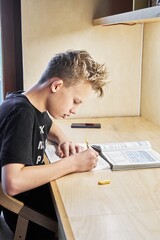 Concentrated teenage boy in black t-shirt does homework writing task in copybook at large wooden table in room close side view