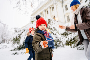 Cute little boy in a knitted red hat holding fireworks on background in a city park. Cute blonde child with dream.Happy kid enjoy the fire sparks. Holidays winter snow childhood concept.