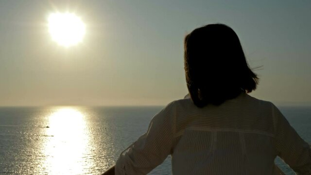 Young woman stands on a balcony with a beautiful view of the sea and mountains. Woman admires the sunset over the sea.