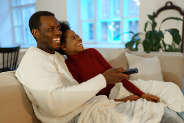 African man and woman relax while watching tv together. Black wife and husband, family, sit on sofa under blanket embracing and happy laughing. Couple in love spend weekend at home. Selective focus