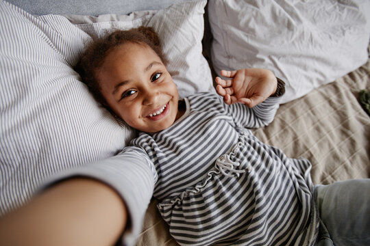 High angle portrait of cute African-American girl taking selfie photo while lying on bed