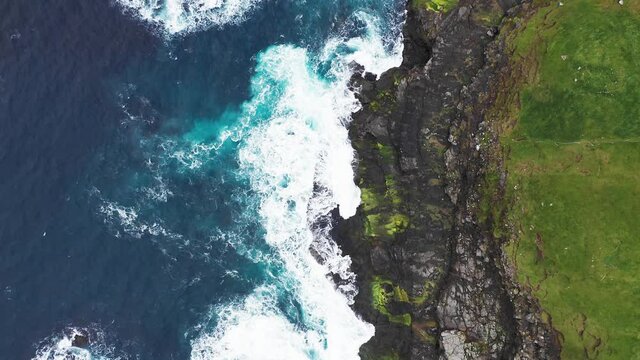 Aerial view of waves break on rocks of Faroe islands cliffs in a blue ocean.Drone Aerial Footage of green nature and the ocean.