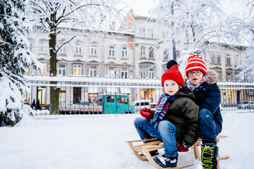 Two different age brothers kids have fun in the beautiful winter park with snow-covered trees. Children walk along a snowy road riding on sled. Winter bright knitted and warm clothes.