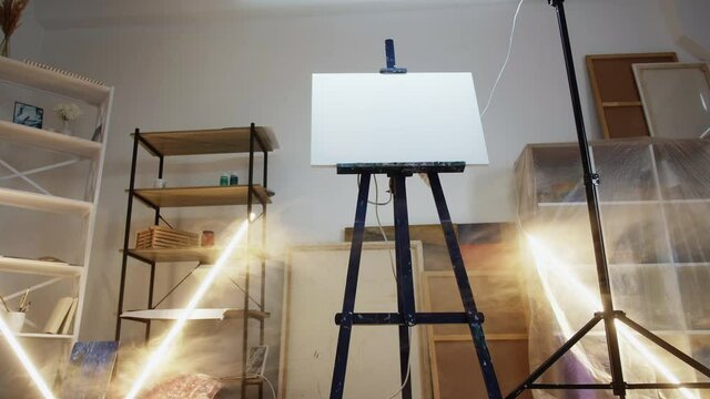 Art studio. Interior scene. Painting equipment. Easel with clear white canvas in middle of light room with artistic tools motion view.