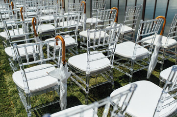 White glass chairs stand on the green grass. Round arch for a wedding ceremony in the background