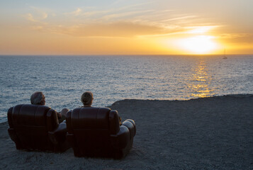 Two elderly people are sitting in armchairs and watching the sunset
