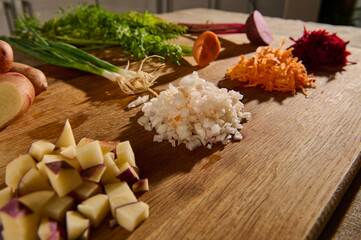 Shredded beet, carrot, onion and cubes of cut potato on a wooden board with halves of vegetables with green leaves and roots on the background. Close-up. Food background