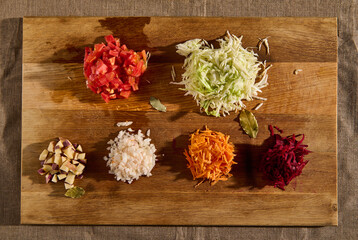 Flat lay of chopped ingredients for a traditional Ukrainian soup- borscht. Chopped and grated raw fresh vegetables -beet, carrot onions, tomatoes, potatoes, and cabbage on a wooden cutting board.