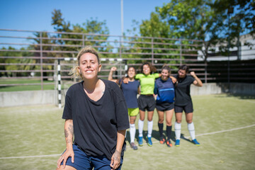 Portrait of young woman on football field. Sportswoman in dark uniform looking proudly at camera, teammates in background. Sport, leisure, female football concept