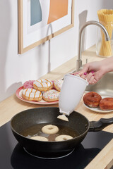 Shot of a female hand that holds a donut mold and pours a liquid dough in a black pan with hot oil. A white donut mold above a pan. Donuts, picture, jar of spaghetti and sink on the background.