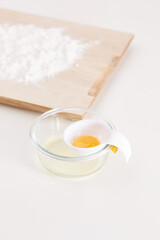 Shot of a transparent glass plate with a white egg separator with silicon holder under the handle. Egg separator with egg on a plate. Wooden cutting board with flour and light table on the background.