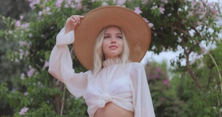 Portrait of romantic stylishly dressed young adult beautiful woman relaxing in a countryside park looking away. Blonde Parisian-styled model in a white transparent dress and straw hat standing against