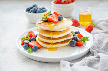 A stack of delicious fluffy pancakes with berries and honey on a white plate on a gray concrete background. Copy space.