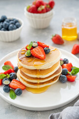 A stack of delicious fluffy pancakes with berries and honey on a white plate on a gray concrete background. Copy space.