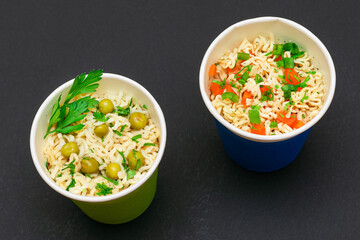 Prepared Instant Noodles with Green Pea, Carrot and Greens in Blue and Green Cups on Black Background. Quick Lunch or Unhealthy Fast Food