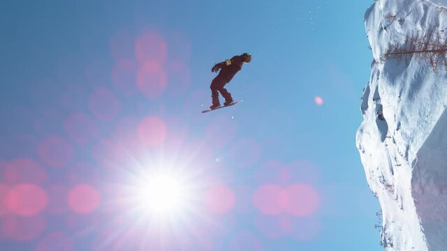 SLOW MOTION, VERTICAL, LENS FLARE: Bright winter sunbeams shine on a professional male snowboarder doing a backflip after taking off a kicker. Action shot of a freestyle snowboarder doing a flip.