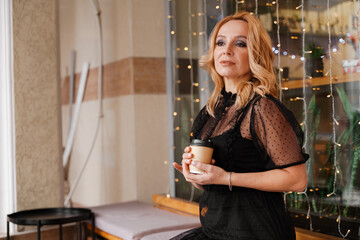 Fototapeta na wymiar Young charming blonde with a cute smile and makeup while relaxing in a cafe. She is holding a cup of coffee in her hands. She is dressed in a black dress with transparent sleeves.