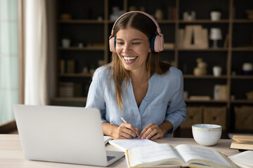 Happy millennial female student in big wireless headphones talking on video call at laptop,...