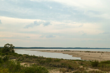 The coastal line of a sandy beach by the Baltic Sea on Sobieszewo Island, Poland, overgrown with high grass and bushes. A small pond next to the shore.The sea is gently waving. A bit of overcast.
