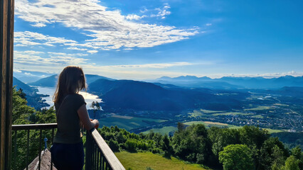 Fototapeta na wymiar A woman standing at the viewing platform of a chapel at the top of Oswaldiberg with the view on the Lake Ossiach in Austrian Alps, Carinthia. The woman enjoys panoramic view. Morning vibe, Freedom