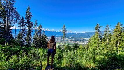 Fototapeta premium A woman standing in a forest in the Austrian Alps, Carinthia. The woman enjoys panoramic view on Villach and the Karawanks mountain chain in the back. Wanderlust, freedom and serenity.