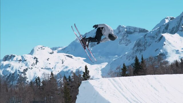 SLOW MOTION: Athletic male skier riding in the sunny Slovenian mountains takes off a kicker and does a rotating grab trick. Action shot of a freestyle skiing pro doing spectacular tricks at snowpark.