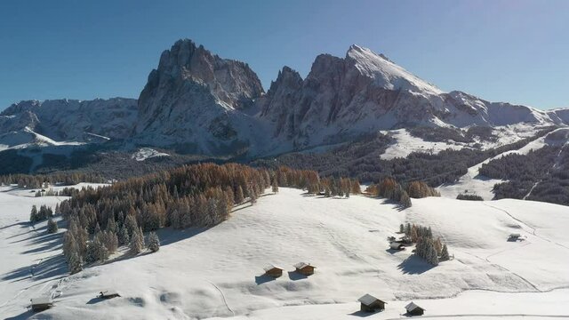 Picturesque landscape with small wooden house, cottage or log cabins on meadow Alpe di Siusi, Seiser Alm, Dolomites, Italy. Snowy hills with orange larch and Sassolungo and Langkofel mountains group