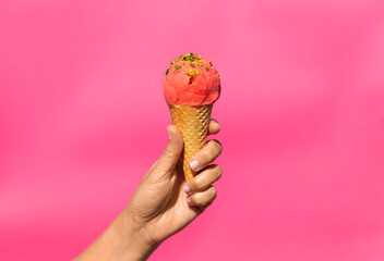 food, dessert and eating concept - close up of hand holding ice cream in waffle cone with pistachio sprinkles over pink background