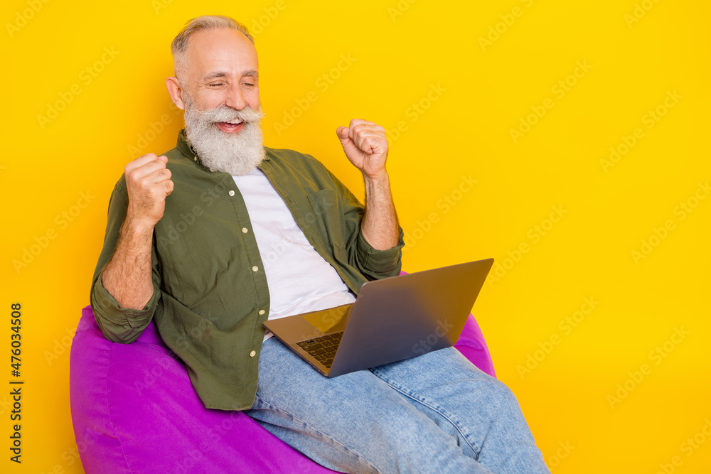 Sticker Photo of astonished man sit beanbag hold laptop look screen raise fists wear green shirt isolated yellow color background