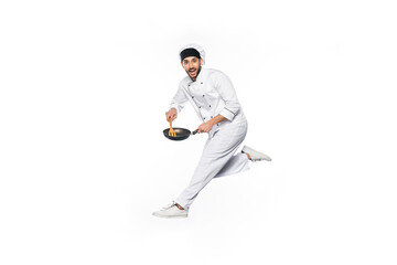 smiling chef in hat and uniform jumping and holding frying pan with wooden spatula isolated on white.