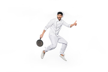 cheerful chef in hat and uniform jumping, showing thumb up while holding frying pan isolated on white.