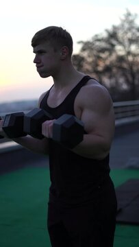 Caucasian white muscular man lifting dumbbells against the sunset sky background. Concept of willpower, motivation and passion. 4k vertical shot 