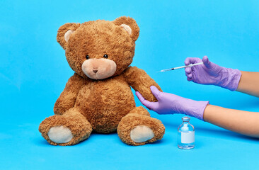 vaccination, healthcare and pandemic concept - hands of doctor in medical gloves with vaccine and syringe making injection teddy bear toy over blue background
