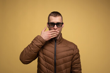 Obraz premium A young man of the 20s in a brown jacket, on a yellow background, sad, wearing glasses for watching a 3d movie covers his mouth with his hand